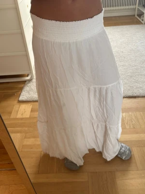 Vit långkjol ! - Säljer en vit långkjol med bred smock i midjan och lätt volang nedtill. Kjolen har en luftig och flowy känsla, perfekt för varmare dagar. Materialet känns mjukt och bekvämt, och kjolen har en enkel men stilren look.