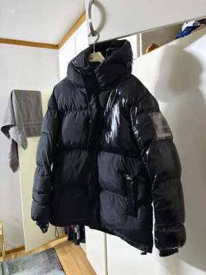 Svart dunjacka från Moose Knuckles - Säljer en svart, glansig dunjacka från Moose Knuckles med stor huva och patch på ärmen. Jackan har dragkedja och tryckknappar framtill, två stora fickor och justerbar nederkant. Perfekt för kalla vinterdagar och riktigt snygg streetstil.