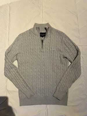 Ljusgrå kabelstickad tröja Bondelid - Kabelstickad ljusgrå tröja från Bondelid med halv dragkedja och ståkrage. Tröjan har långa ärmar, ribbade muddar och klassisk design. Perfekt för lager-på-lager och snygg till jeans eller chinos.