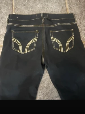 Svarta jeans från Hollister - Snygga svarta jeans från Hollister med kontrasterande beiga sömmar och coola bakfickor med unikt mönster. Klassisk femficksmodell och knappstängning framtill. Perfekta för en trendig och avslappnad stil.