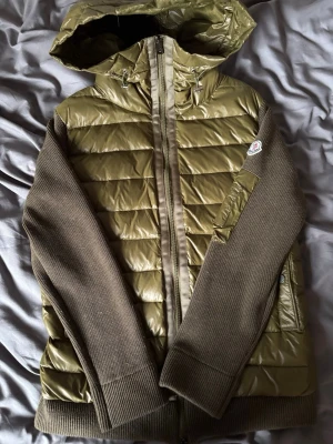 Moncler cardigan Olivgrön  - Snygg olivgrön dunjacka från Moncler med glansigt quiltat tyg och stickade ärmar i mörkare grön nyans. Jackan har huva, dragkedja framtill, två fickor med tryckknappar och Moncler-logga på ärmen. Perfekt för kalla vinterdagar.