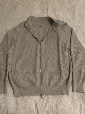 Ljusgrå ribbad ziptröja - Ljusgrå ribbad ziptröja med hög krage och två fickor framtill. Tröjan har långa ärmar och breda muddar vid ärmslut och nederkant. Perfekt för lager på lager och enkel att matcha med olika outfits.