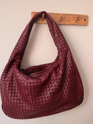 Vinröd flätad axelväska i läder - Snygg vinröd handväska med flätad design och mjuk, rundad form. Väskan har ett bredare axelband och dragkedja upptill. Tillverkad i läder och perfekt för dig som gillar en unik och trendig look.