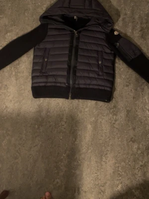 Svart vindjacka med huva - Svart vindjacka med quiltad kropp och stickade ärmar. Jackan har dragkedja framtill, två fickor med knappar och en extra ficka på ena ärmen. Huva och ribbade muddar ger en sportig look. Perfekt för blåsiga dagar.