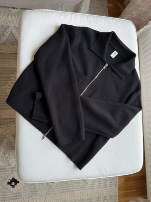 Svart ziptröja från Åhléns - Snygg svart ziptröja från Åhléns med krage och dragkedja framtill. Tröjan har en minimalistisk design och är tillverkad i ett mjukt, tjockare material som känns lyxigt. Perfekt för dig som gillar stilrena plagg med modern känsla.