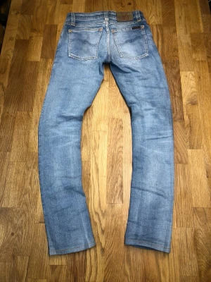 Ljusblå jeans från Nudie Jeans - Säljer ett par ljusblå jeans från Nudie Jeans med klassisk femficksmodell och snygga slitningar. Jeansen har raka ben och kontrastsömmar samt en läderpatch med logga bak i midjan. Tillverkade i bomull för en skön och avslappnad look.