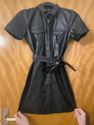 Svart kort klänning i skinnimitation - OBS INTE MINA BILDER! Köpte från Vinted för 150kr  men passade mig inte som jag ville❤️Snygg svart kort klänning i skinnimitation med knappar framtill, korta ärmar och bälte i midjan. Två bröstfickor med lock ger en cool touch. Perfekt för dig som vill sticka ut med en edgy look.