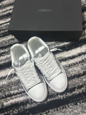 Vita och grå sneakers från Numeris - Stilrena sneakers från Numeris i vitt och grått med coolt camomönster på plösen. Skorna har klassisk låg profil, breda vita snören och är tillverkade i slätt material med detaljer i textil. Perfekta för dig som gillar en clean och modern look.