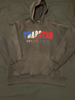 Trapstar hoodie med broderad text - Säljer en mörkgrå hoodie från Trapstar med broderad text i blått, vitt och rött på bröstet där det står 'TRAPSTAR' och 'IT'S A SECRET' spegelvänt. Klassisk känguruficka och huva. Perfekt för dig som gillar streetwear och vill sticka ut. Oops Den är lite sliten och men inte stora skador utan jätte små inte ens synliga :)