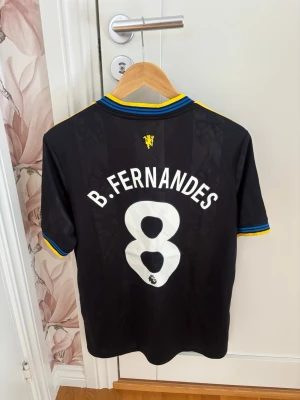 Manchester United B. Fernandes tröja - Snygg svart Manchester United matchtröja med gula och blå detaljer, nummer 8 och B. Fernandes på ryggen. Tröjan har klubbmärke, Adidas-logga och Snapdragon sponsortryck framtill. Perfekt för fotbollsfans som vill sticka ut på läktaren eller planen. Nyskick, tredjeställ 25/26 och storlek M