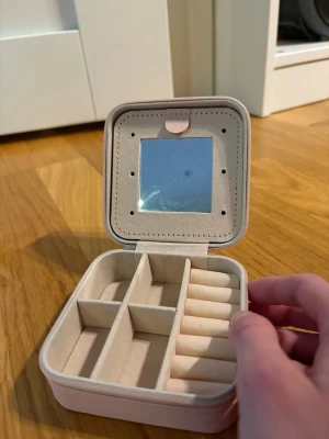 Ljusrosa smyckesask med spegel - Praktisk och stilren ljusrosa smyckesask i fyrkantig form med spegel på insidan av locket. Flera fack för ringar, örhängen och halsband samt elastisk ficka och krokar för extra förvaring. Tillverkad i mjukt material med sammetsliknande yta.