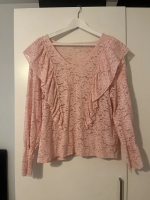 Rosa spetsblus med volanger - Söt rosa blus i spets med långa ärmar och volangdetaljer framtill. Blusen har v-ringning och ett romantiskt uttryck tack vare det genombrutna spetsmönstret. Perfekt för dig som gillar en feminin och trendig stil.