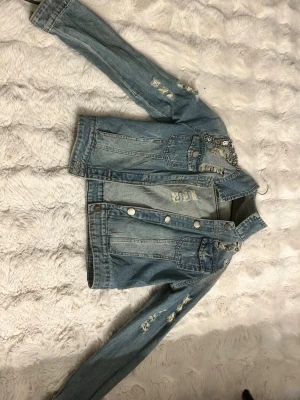 Ljusblå distressed jeansjacka - Trendig ljusblå jeansjacka med slitna detaljer och silverfärgade knappar. Klassisk modell med två bröstfickor och dekorativa sömmar. Snygg kedjedetalj vid axeln som ger extra edge till looken. Perfekt för dig som vill ha en cool och avslappnad stil.