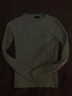 Ljusgrå kabelstickad tröja Gant - Snygg ljusgrå kabelstickad tröja från Gant i stretchig bomull. Tröjan har rund halsringning, långa ärmar och diskret Gant-logga på bröstet. Perfekt basplagg med ribbade muddar vid ärmslut och nederkant.