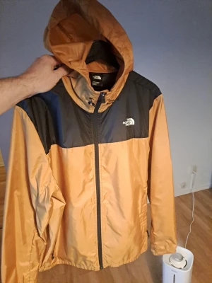 North Face brun och svart vindjacka - Den ursprungliga Mountain Jacket blev en ikon av goda skäl. Utvecklad 1985 för att ge klättrare samma skydd mot extrema förhållanden som skidåkare, har den värmt ryggen på några av våra största bergsatleter på expeditioner över hela världen. Säsongens version har elastisk huva, fåll och manschetter för fullständig komfort och flexibilitet. Och när temperaturen stiger kan den packas ner i sin egen ficka, redo att användas igen när du behöver den nästa gång. Lämna inte hemmet utan den.