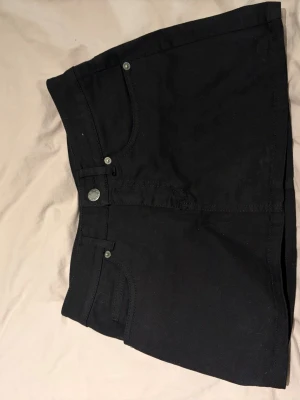 Svart kort jeanskjol - Säljer en svart kort jeanskjol med klassisk femficksdesign och bälteshällor. Kjolen har knapp- och dragkedjestängning framtill samt raka sömmar. Perfekt basplagg till garderoben! Storlek 32 men motsvara ungefär xxs-xs.