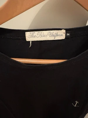 Svart t-shirt från The Blue Uniform - En svart t-shirt från The Blue Uniform med en liten vit ankare-brodyr på bröstet. Klassisk rund hals och korta ärmar. Tillverkad i mjuk bomull som känns skön mot huden. Enkel och stilren design som passar till det mesta.