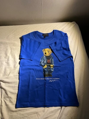 Blå Polo Bear t-shirt från Ralph Lauren - Snygg blå t-shirt från Polo Ralph Lauren med den ikoniska Polo Bear-trycket på framsidan. Klassisk rund hals och korta ärmar. Tillverkad i mjuk bomull som känns skön mot huden. Perfekt för dig som vill ha en stilren men lekfull look. Nypris kostar den 1149. Om du har några funderingar har vänlig och ställ frågor.