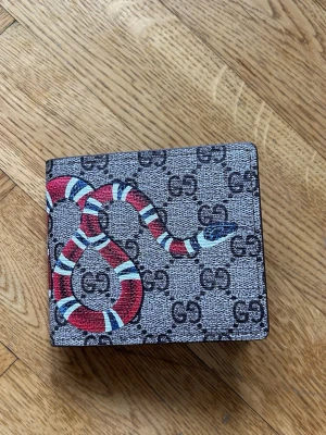 Gucci plånbok med ormmotiv - Snygg plånbok från Gucci i canvas med klassiskt GG-mönster och en röd, vit och blå orm tryckt över hela utsidan. Plånboken har en fyrkantig form och är perfekt för dig som vill sticka ut med en trendig accessoar.