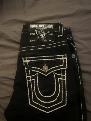 Svarta True Religion jeans  - Säljer ett par svarta jeans från True Religion med tydliga vita kontrastsömmar och stor fickdetalj bak. Klassisk femficksmodell med unika detaljer och logga på bakfickan. Perfekta för dig som vill sticka ut med din denimstil.