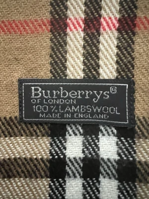 Beige rutig halsduk från Burberrys - Klassisk beige halsduk från Burberrys i 100% lammull med ikoniskt rutmönster i svart, vitt och rött. Halsduken har fransar i ändarna och är tillverkad i England. Perfekt accessoar för att lyfta din outfit med en tidlös look.