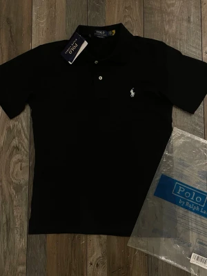 Svart pikétröja från Ralph Lauren - Klassisk svart pikétröja från Ralph Lauren med vit broderad logga på bröstet. Tröjan har krage, knappar framtill och är tillverkad i bomull. Perfekt för en stilren och tidlös look.