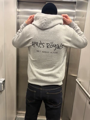 Grå hoodie Après Royale  - Snygg grå hoodie från Après Royale med svart tryck på ryggen där det står 'APRÈS ROYALE' och 'NOLT NORDIC ALPINE'. Hoodien har en klassisk huva och är tillverkad i mjukt material, perfekt för en avslappnad stil.