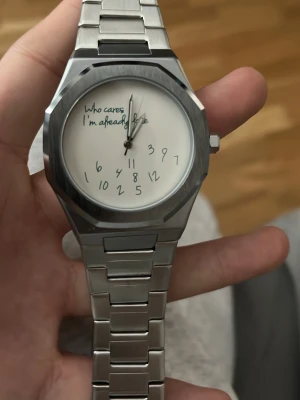 Silverfärgad klocka från Miler - Unik klocka från Miler i silverfärgat rostfritt stål med vit urtavla. Urtavlan har handskrivna siffror och texten 'Who cares I'm already late' för en lekfull touch. Klockan har länkarmband och modern, kantig form.