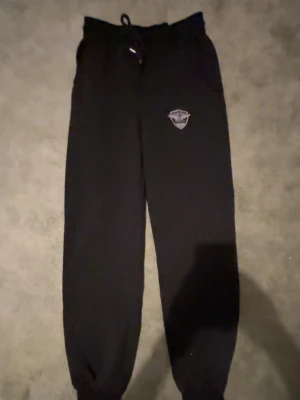 Svarta mjukisbyxor med tryck - Säljer ett par svarta mjukisbyxor med resår i midjan och benslut samt snörning i midjan. Byxorna har ett vitt tryck på ena benet med texten 'Athletic Dept'. Perfekta för chill dagar eller träning. Klassisk passform och mjukt material.