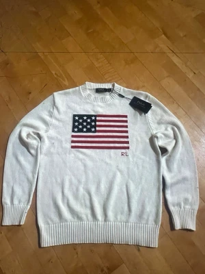 Vit stickad tröja Polo Ralph Lauren - Vit stickad tröja från Polo Ralph Lauren med amerikansk flagga broderad på bröstet och RL-broderi. Tröjan har rund halsringning och ribbstickade muddar vid ärmslut och nederkant. Perfekt för dig som gillar klassisk och stilren design. Storlek L men passar mer som M