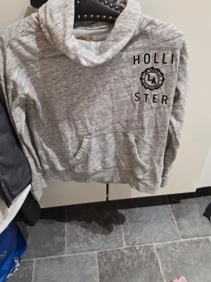 Grå hoodie från Hollister LA - Snygg gråmelerad hoodie från Hollister med tryck 'HOLLISTER LA' på bröstet. Hoodien har en stor känguruficka framtill och en mysig huva. Perfekt för chill dagar och streetwear-stil.
