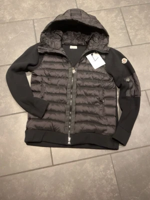 Svart vindjacka med huva från Moncler - Snygg svart vindjacka från Moncler med quiltad front, stickade ärmar och huva. Jackan har dragkedja framtill, två sidofickor med dragkedja och Moncler-logga på ärmen. Materialmix med dunfylld kropp och ribbade muddar ger en modern look.
