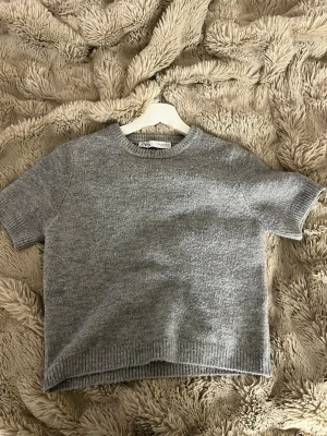 Grå stickad t-shirt från Zara - Kortärmad stickad t-shirt från Zara i mjuk grå ullmix. Tröjan har rund halsringning och ribbade kanter vid ärmslut och nederkant. Enkel och stilren design som passar perfekt till jeans eller kjol.