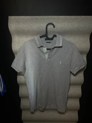 Grå pikétröja från Ralph Lauren - Klassisk grå pikétröja från Ralph Lauren med broderad logga på bröstet. Tröjan har korta ärmar, krage och knappar framtill. Perfekt för en stilren och avslappnad look.