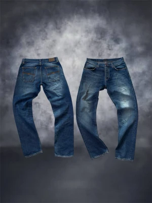 Blå jeans från Nudie jeans steady Eddie  - Snygga blå jeans från Nudie Jeans med klassisk femficksdesign och raka ben. Jeansen har slitningar framtill för en cool, använd look och är gjorda i bomull. Perfekta för dig som gillar en avslappnad stil med edge. Modellen heter steady Eddie litet hål i en av fickorna men inget som märks 