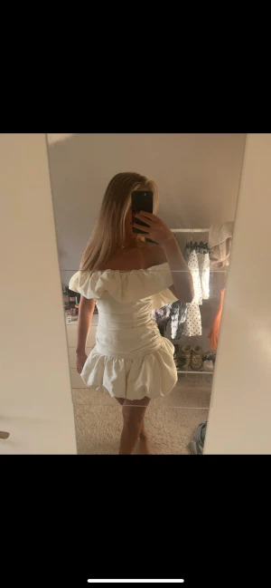 Vit offshoulder volangklänning - Supersöt vit kortklänning med offshoulder och stora volanger både upptill och nertill. Klänningen har en figurnära passform och är perfekt för dig som vill sticka ut. Materialet känns lätt och somrigt, och modellen ger en riktigt trendig vibe.
