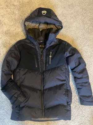 Mörkblå Sail Racing dunjacka - Snygg mörkblå dunjacka från Sail Racing med huva och logga på bröstet. Jackan har två bröstfickor med dragkedja, patch på ärmen och justerbara ärmslut. Perfekt för kalla dagar med sin puffiga design och sportiga look.