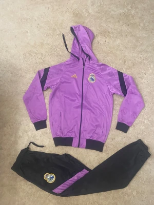 Lila Real Madrid vindjacka Adidas - Säljer en lila vindjacka från Adidas med Real Madrid-logga på bröstet och guldiga Adidas-detaljer. Jackan har huva med snörning, svarta muddar och dragkedja framtill. Materialet är glansigt polyester och jackan har svarta detaljer på ärmarna.