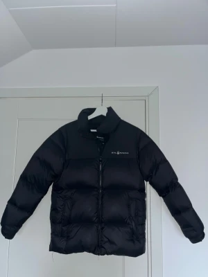 Svart pufferjacka från Sail Racing - Svart pufferjacka från Sail Racing med hög krage och diskret logga på bröstet. Jackan är fylld med dun och fjäder (600 fill power) och har en stilren design med dragkedja framtill. Materialet är syntet och dun, perfekt för kalla dagar.