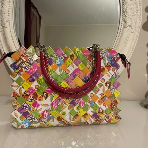 Färgglad handväska av godispapper - Unik handgjord handväska flätad av färgglada godispapper med rosa handtag  och dragkedja upptill. Väskan har ett lekfullt rutigt mönster i flera färger och är perfektiögonfallande accessoar.