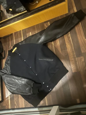 Svart bomberjacka med skinnärmar - Säljer en svart bomberjacka med snygga svarta skinnärmar och knappar framtill. Jackan har en klassisk college-look med ribbade muddar och en diskret ficka med dragkedja på bröstet. Perfekt för dig som gillar stilren och cool streetwear.