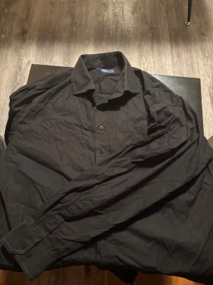 Svart skjorta från Oscar of Sweden - Svart skjorta från Oscar of Sweden i storlek 46. Klassisk modell med knappar framtill, lång ärm och diskreta vita sömmar på krage och manschetter. Skjortan är gjord i mjuk bomull och har en stilren look som funkar till många olika outfits.