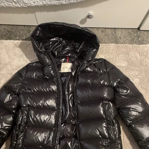 Svart Moncler jacka - Säljer en svart pufferjacka från Moncler med glansig finish och huva. Jackan har dragkedja framtill, två sidofickor och coolt Moncler-band på insidan. Insidan har ett unikt serietidningsmotiv. Perfekt för dig som vill ha en varm och snygg jacka med street vibe. Från NK och köpt för 5000kr