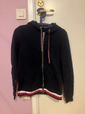Marinblå hoodie från Tommy Hilfiger - Snygg marinblå hoodie från Tommy Hilfiger med dragkedja, huva och coola snören i rött, vitt och blått. Tröjan har ribbad kant med röd och vit rand längst ner och praktiska fickor framtill. Perfekt för chill dagar och streetstyle.