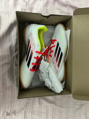 Adidas F50 Club IN fotbollsskor - Säljer ett par Adidas F50 Club IN fotbollsskor i vitt med svarta och röda detaljer, neongul insida och röda snören. Skorna har låg profil och platt sula, perfekta för inomhusspel. Tillverkade i syntetmaterial för lätt känsla och bra bollkontroll.