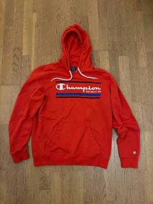 Röd Champion hoodie med tryck - Säljer en röd hoodie från Champion i storlek medium. Tröjan har vit snörning vid huvan, stor logga och tryck på bröstet samt en klassisk magficka. Materialet är mjuk bomull och polyester, perfekt för chill dagar. Ribbade muddar vid ärmslut och nederkant.