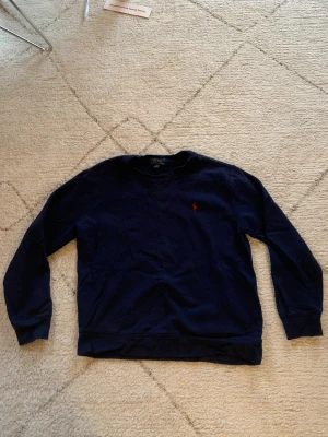 Mörkblå sweatshirt från Polo Ralph Lauren - Säljer en mörkblå sweatshirt från Polo Ralph Lauren med klassisk röd logobrodering på bröstet. Tröjan har rund hals, långa ärmar och ribbade muddar. Materialet är mjuk bomull som är skönt mot huden och passar perfekt till chill dagar.