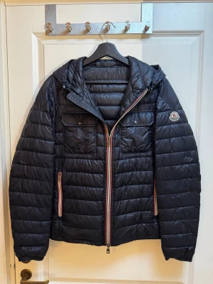 Svart Moncler dunjacka med huva - Svart dunjacka från Moncler med huva, två bröstfickor med knappar och snygga detaljer i rött och vitt vid dragkedjorna. Jackan har quiltad design, Moncler-logga på ärmen och dubbla dragkedjor framtill. Perfekt för kalla dagar och riktigt stilren. Drag kedjan har gått sönder men funkar fortfarande. Den har 2 lagningar en på ryggen och en på framsidan. Dom syns inte när man har på sig jackan. Jackan har stl 2 