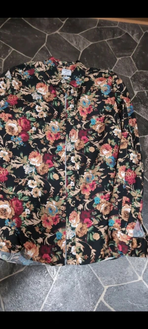 Blommig långärmad skjorta stl L - Säljer en riktigt snygg långärmad skjorta med blommigt mönster i färger som beige, röd, blå och grön mot svart bakgrund. Skjortan har klassisk krage och knappar framtill. Perfekt för dig som gillar färgstarka och unika plagg.