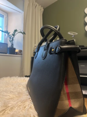 Fin svart väska  - Säljer en snygg svart handväska med rutiga sidor. Köpt på Vinted för några år sedan men har inte blivit använd, så nu får den vandra vidare. Stadig modell med både handtag och avtagbar axelrem. Gott om plats inuti och en dragkedjeficka. Fint skick. Inspirerad av märket Burberry  